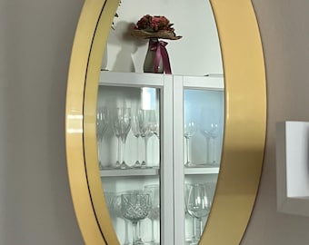 Könnte beinhalten: Ein großer, ovaler Spiegel mit einem dicken, cremefarbenen Rahmen. Der Spiegel spiegelt einen weißen Schrank mit Glasböden und eine Vase mit rosa Blumen wider.