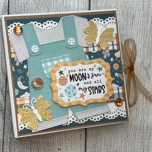May include: A square scrapbook album featuring a teal paper overall cutout, two yellow butterfly die-cuts, and amber rhinestones. The center label reads: you are my MOON & sun and all my STARS. The background has a celestial pattern and a brown ribbon tie.