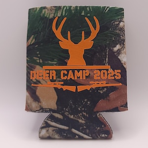 May include: A camouflage-patterned can cooler with orange text and graphics. The text reads "DEER CAMP 2025" with a deer silhouette and two rifle silhouettes. The cooler is designed to keep beverages cold.