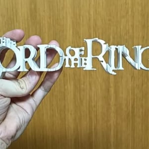 May include: A white, three-dimensional sign reading "THE LORD OF THE RINGS" is held in a hand. The sign is made of a light-coloured material and is against a wooden background. The sign is a decorative item.