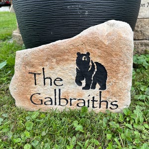 May include: A tan-colored natural stone garden marker featuring a black silhouette of a bear and the engraved black text "The Galbraiths". The stone has an irregular, jagged edge and a textured surface, positioned horizontally on the ground.