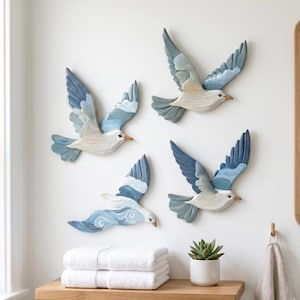 Puede incluir: Un conjunto de cuatro esculturas de gaviotas de madera para pared, cada una con detalles pintados a mano en tonos azul, blanco y gris. Las aves se muestran en varias posiciones de vuelo con las alas extendidas y texturas de plumas talladas.