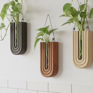 Peut inclure: Trois jardinières murales suspendues, de couleurs grise, marron et bois naturel. Chaque jardinière présente un design en forme de U avec des rainures verticales et un tube en verre pour les plantes. Des plantes vertes à feuilles sont affichées.