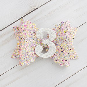 Op de afbeelding: Een glitter haarstrik in pastelkleuren met een zilveren nummer 3 in het midden. De strik is versierd met roze, gele en paarse glitter. De strik ligt op een wit houten oppervlak.