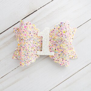 Op de afbeelding: Een pastelkleurige glitterstrik met een wit cijfer 1 in het midden. De strik is versierd met een verscheidenheid aan pastelkleuren, waaronder roze, geel en blauw. Het cijfer 1 is gemaakt van witte glitter.