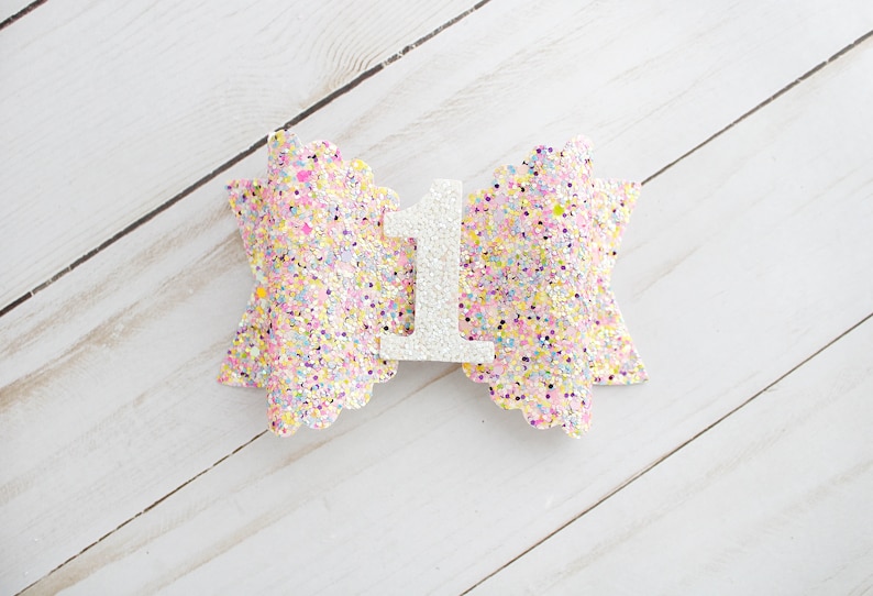Op de afbeelding: Een pastelkleurige glitterstrik met een wit cijfer 1 in het midden. De strik is versierd met een verscheidenheid aan pastelkleuren, waaronder roze, geel en blauw. Het cijfer 1 is gemaakt van witte glitter.