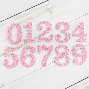 May include: Pink glitter numbers 0 to 9 on a white wood background.