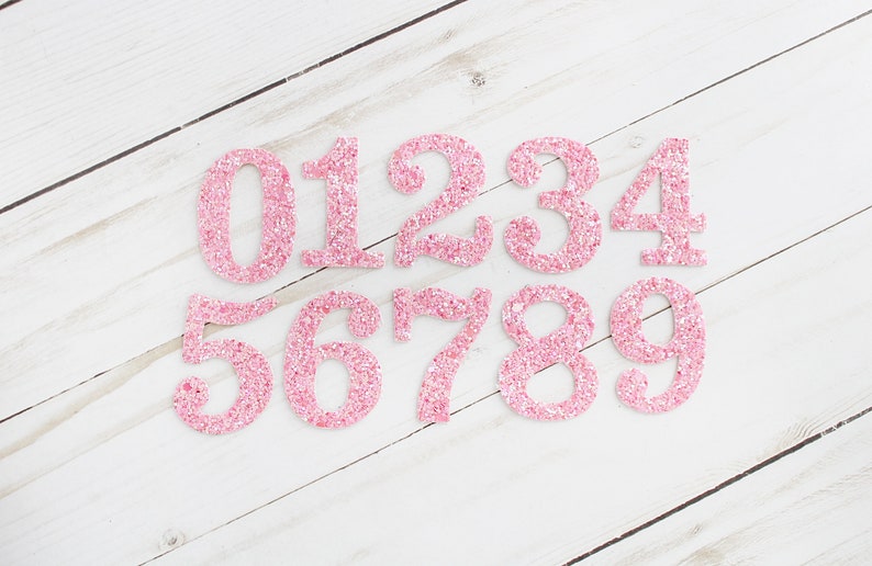May include: Pink glitter numbers 0 to 9 on a white wood background.