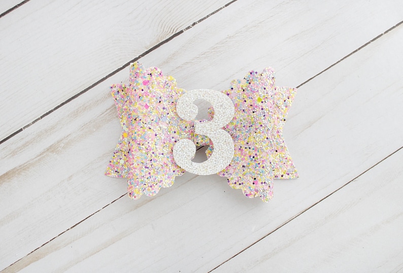 Op de afbeelding: Een glitter haarstrik in pastelkleuren met een zilveren nummer 3 in het midden. De strik is versierd met roze, gele en paarse glitter. De strik ligt op een wit houten oppervlak.