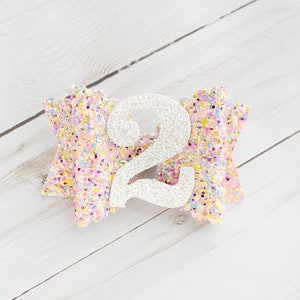 Op de afbeelding: Een glitter haarstrik in pastelkleuren met een groot wit cijfer 2 in het midden. De strik is versierd met een confetti-achtig patroon en staat op een witte houten achtergrond. Dit accessoire is perfect voor een tweede verjaardag.