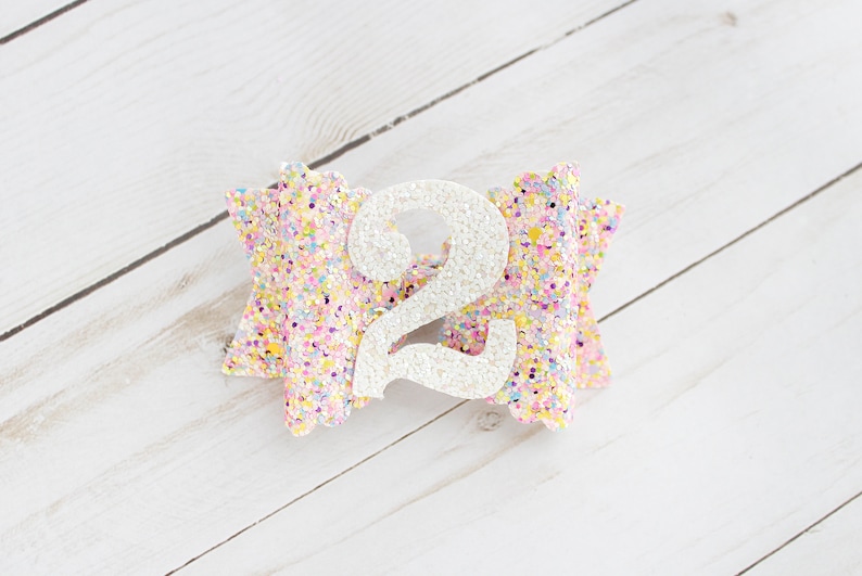 Op de afbeelding: Een glitter haarstrik in pastelkleuren met een groot wit cijfer 2 in het midden. De strik is versierd met een confetti-achtig patroon en staat op een witte houten achtergrond. Dit accessoire is perfect voor een tweede verjaardag.