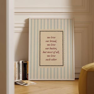 May include: A framed print with a cream-colored background and vertical blue stripes. The print features the text "we love our bread, we love our butter, but most of all, we love each other" in a decorative font. Books and a yellow ottoman are in the background.
