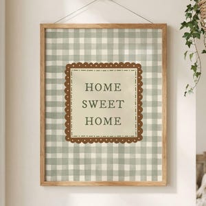 Puede incluir: Una obra de arte enmarcada con un marco de madera marrón claro. La obra presenta un fondo a cuadros verdes y blancos con un borde festoneado marrón alrededor del texto "HOME SWEET HOME". El arte está colgado en una pared.