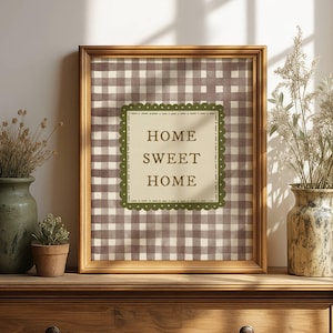 Puede incluir: Un cuadro enmarcado con el texto "HOME SWEET HOME" sobre un fondo a cuadros marrón y blanco. El marco dorado está sobre una cómoda de madera. Dos jarrones y una pequeña planta en maceta completan la escena.