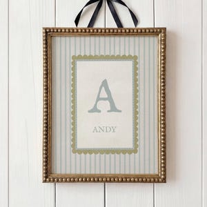 May include: Framed wall art featuring a light blue letter "A" and the name "ANDY" in a decorative frame. The artwork has a striped background with a scalloped border and is accented with a black ribbon.