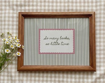 May include: A rectangular wooden frame containing a light-colored background with thin vertical stripes. Centered is a white rectangular card with a scalloped pink border featuring the black cursive text: So many books, so little time.