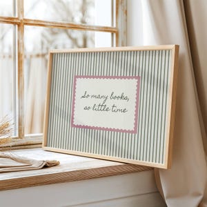 May include: A rectangular framed print featuring a light green and white vertical striped background. In the center, a cream-colored rectangular label with a pink scalloped border displays the black text: So many books, so little time.