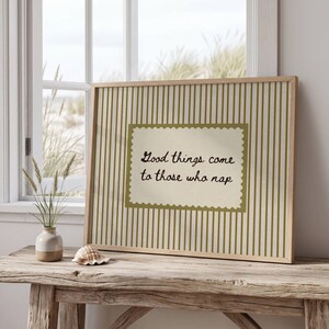 May include: A framed print with the text "Good things come to those who nap." The artwork features a light wooden frame, a striped background, and a decorative border around the text. The print is displayed on a rustic wooden table.