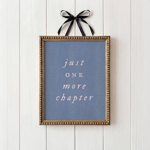 May include: A rectangular wall sign featuring a blue background with the white text "just ONE more chapter" centered in the middle. The sign is enclosed in a beaded gold frame and is suspended by a black ribbon tied in a bow at the top.