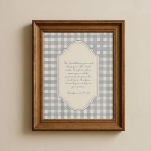 May include: A framed print featuring a light blue and white gingham pattern background. The print has a central cream-colored, ornate shape with script text from Numbers 6:24-26. The frame is a warm brown color.