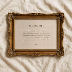 Puede incluir: Marco dorado ornamentado con un papel crema en el interior. El texto en el papel dice "THE BLESSING" seguido de una oración. El marco tiene detalles de volutas y una estética vintage.