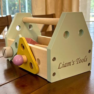 May include: A wooden toy toolbox in light green and natural wood tones, featuring a handle and the engraved text Liam's Tools. The box includes a yellow triangular piece, a purple knob, and various holes for assembly, showcasing a multi-colored design.