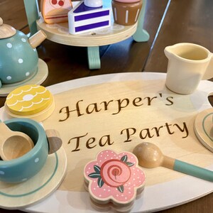 May include: Wooden toy tea set with a teal teapot, cups, and a serving tray. The tray is engraved with "Harper's Tea Party." The set includes a tiered stand with wooden desserts. The tea cups and teapot have white polka dots.