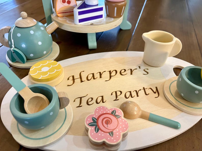 May include: Wooden toy tea set with a teal teapot, cups, and a serving tray. The tray is engraved with "Harper's Tea Party." The set includes a tiered stand with wooden desserts. The tea cups and teapot have white polka dots.