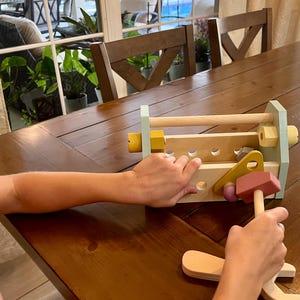 May include: A wooden toy workbench featuring a light blue frame, natural wood surfaces, and various holes for assembly. The set includes a pink hammer, yellow and light blue nuts, and bolts, designed for interactive construction play and fine motor skill development.