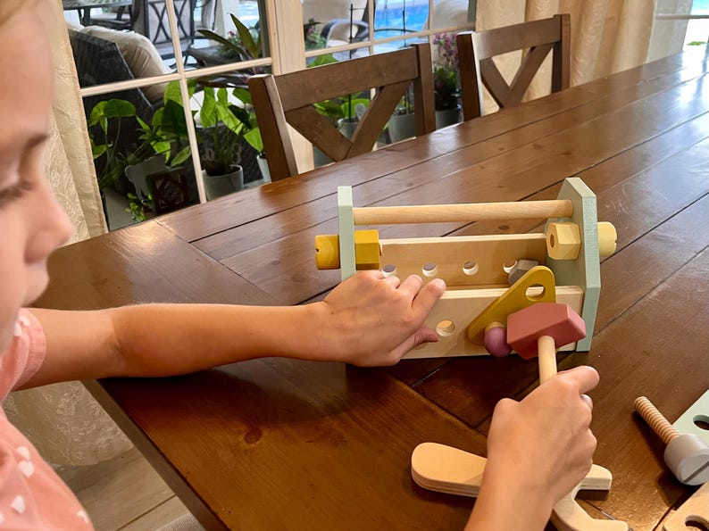 May include: A wooden toy workbench featuring a light blue frame, natural wood surfaces, and various holes for assembly. The set includes a pink hammer, yellow and light blue nuts, and bolts, designed for interactive construction play and fine motor skill development.