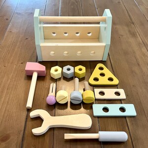 May include: A wooden toy tool set featuring a light blue and natural wood toolbox with holes, a pink hammer, a wrench, a screwdriver with a light blue handle, three hexagonal nuts, three bolts, and three flat wooden construction pieces in pastel colors.