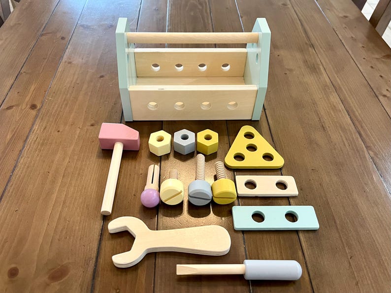 May include: A wooden toy tool set featuring a light blue and natural wood toolbox with holes, a pink hammer, a wrench, a screwdriver with a light blue handle, three hexagonal nuts, three bolts, and three flat wooden construction pieces in pastel colors.