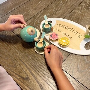 May include: A wooden tea set with a teal teapot, cups, saucers, spoons, and a serving tray. The tray has the text "Isabella's Tea Party" written on it, along with wooden cookies and a teacup with a lemon slice.