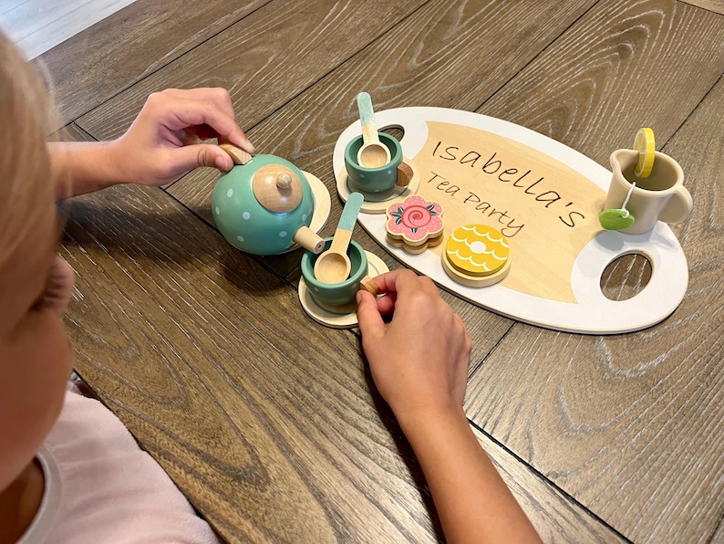May include: A wooden tea set with a teal teapot, cups, saucers, spoons, and a serving tray. The tray has the text "Isabella's Tea Party" written on it, along with wooden cookies and a teacup with a lemon slice.