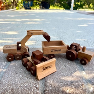 May include: Wooden toy construction vehicles, including an excavator, a bulldozer, and a tractor with a trailer. The excavator and bulldozer have names engraved on them. The toys are made of light and dark brown wood.