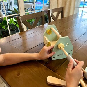May include: A light green wooden toy toolbox with the text Liam's Tools printed on the side. The set includes a wooden handle, yellow and light green bolts, and various wooden construction pieces with holes, designed for assembly and play.