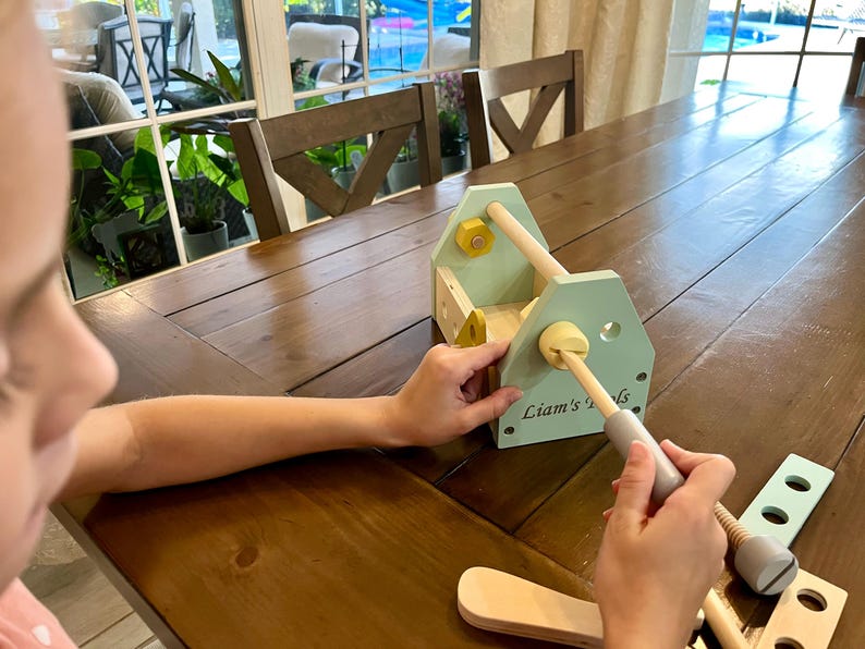 May include: A light green wooden toy toolbox with the text Liam's Tools printed on the side. The set includes a wooden handle, yellow and light green bolts, and various wooden construction pieces with holes, designed for assembly and play.