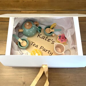 May include: A wooden tea set in a white box. The set includes a teal teapot with white polka dots, cups, spoons, a creamer, and a flower-shaped cookie. The words "Kate's Tea Party" are on a wooden sign.