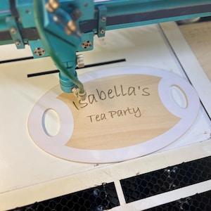 May include: A laser engraver etches the words "Isabella's Tea Party" onto a wooden oval sign with white trim. The sign is on a white surface, and the laser machine is teal.