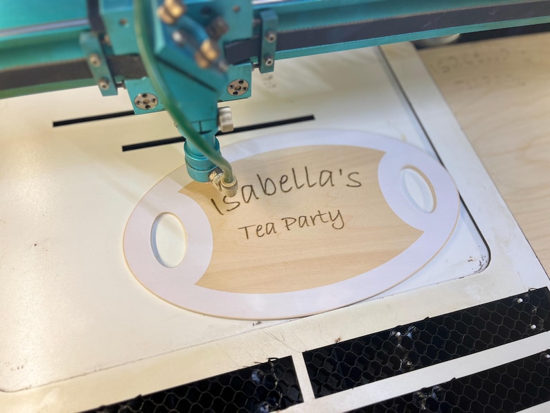 May include: A laser engraver etches the words "Isabella's Tea Party" onto a wooden oval sign with white trim. The sign is on a white surface, and the laser machine is teal.