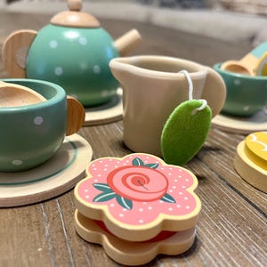 May include: A wooden toy tea set with a green teapot and cups, a creamer, and a flower-shaped sandwich. The tea set includes a wooden spoon, a lemon slice, and a tea bag. The tea set is painted with white polka dots.