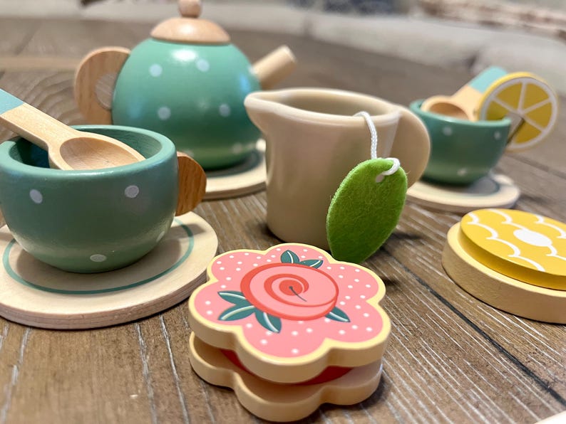 May include: A wooden toy tea set with a green teapot and cups, a creamer, and a flower-shaped sandwich. The tea set includes a wooden spoon, a lemon slice, and a tea bag. The tea set is painted with white polka dots.