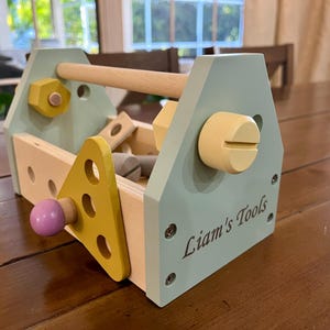May include: A wooden toy toolbox in light blue, yellow, and natural wood tones. The side panel is engraved with the text Liam's Tools. It features a handle, various holes, and attached wooden components including a yellow triangle and a purple knob.