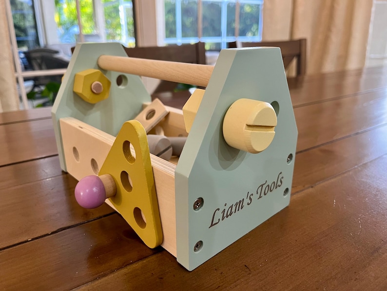 May include: A wooden toy toolbox in light blue, yellow, and natural wood tones. The side panel is engraved with the text Liam's Tools. It features a handle, various holes, and attached wooden components including a yellow triangle and a purple knob.