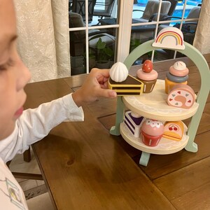 May include: A wooden toy dessert stand with a rainbow arch and two tiers. The stand holds various wooden desserts, including a slice of cake, cupcakes, and a roll cake. The desserts are in pastel colors. A hand is holding a piece of cake.