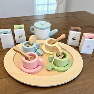Puede incluir: Un juego de té de madera con una tetera, cuatro tazas y platillos, y cuatro recipientes para condimentos etiquetados como "Azúcar", "Té", "Café" y "Miel". El juego está dispuesto en una bandeja de madera.