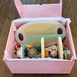 May include: A pink box filled with wooden toy tea set pieces. The set includes a wooden serving tray, a teapot, cups, and a rainbow-colored cake. The toys are in various colors, including green, pink, and natural wood tones.