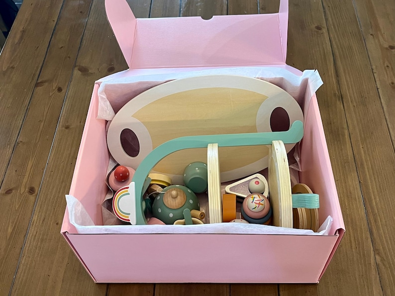 May include: A pink box filled with wooden toy tea set pieces. The set includes a wooden serving tray, a teapot, cups, and a rainbow-colored cake. The toys are in various colors, including green, pink, and natural wood tones.