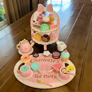 May include: A wooden tea party set featuring a pink tiered stand, teapot, cups, and toy desserts. The set includes a tray with the text "Charlotte's Tea Party" and decorative elements, perfect for imaginative play.