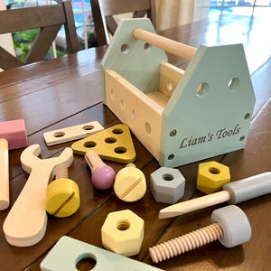 May include: A wooden toy tool set with a light blue toolbox and various tools. The toolbox has a wooden handle and the text "Liam's Tools". The set includes a pink hammer, a yellow wrench, and other colorful wooden pieces.